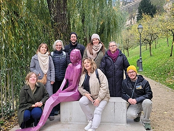 Acht Personen des Fröbel-Kindergartens Farbkleckse posieren lächelnd um die violette Statue der sagenhaften Melusina, die auf einer Bank im Stadtteil Grund in Luxemburg sitzt. Im Hintergrund sind herbstliche Bäume, eine Laterne und eine historische Mauer zu sehen.